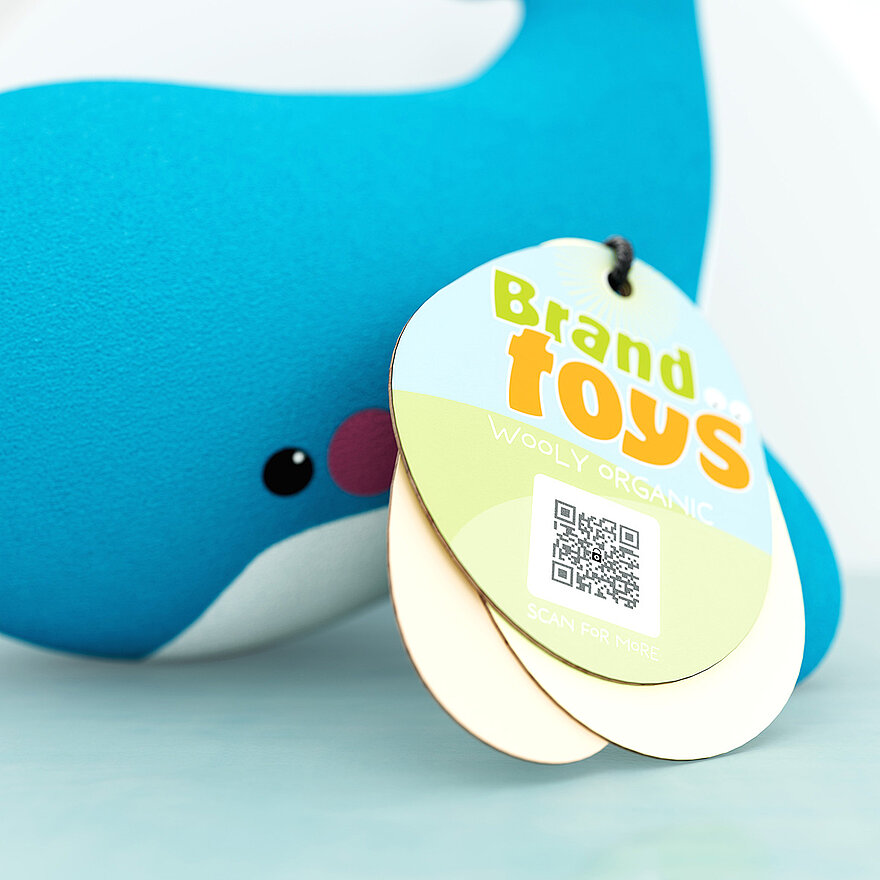 A close-up of a soft blue plush whale toy with a hangtag attached. The hangtag displays the text “Brand Toys Woolly Organic” and features a QR code that incorporates the ValiGate® solution. This QR code includes an embedded security pattern, ensuring advanced product authenticity and counterfeit protection. Printed directly on the hangtag, the ValiGate® QR code offers eco-friendly, seamless verification without additional labels or materials. The tag invites users to “Scan for More,” connecting them to secure product details and demonstrating the innovative integration of anti-counterfeiting technology.