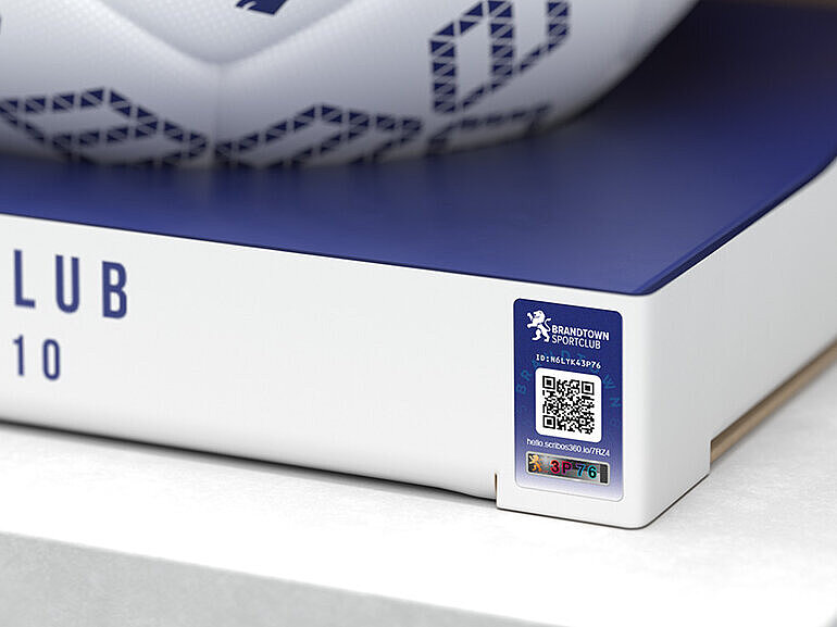 A white soccer ball with blue geometric designs and a lion emblem displayed in open packaging labeled “Sportsclub Since 1910.” The soccer ball features VeoMark® technology, which integrates overt and covert security features for advanced counterfeit protection. This solution ensures product authenticity and enhances brand trust.